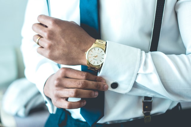 Man in white shirt, blue tie, and black suspenders holding his shirt cuff with black cufflink, gold watch, and gold ring.